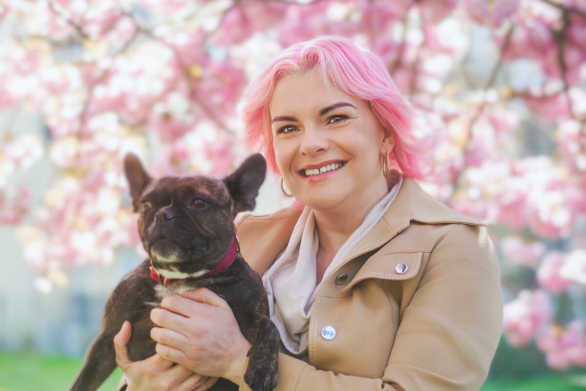 French Bulldog with Frenchie Mom - a picture of a woman with pink hair in a beige jacket holding a french bulldog